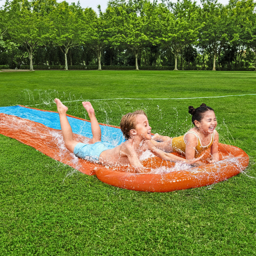 Two children sliding down a inflatable water slide on grass with trees in the background.